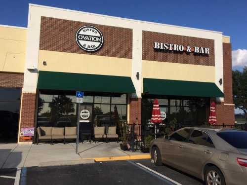 Exterior of a bistro and bar with green awnings, outdoor seating, and a parked car in front.