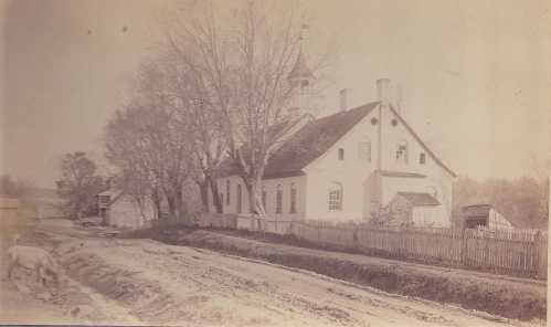 A vintage photo of a rural scene featuring a white building, trees, and a dirt road with a fence.