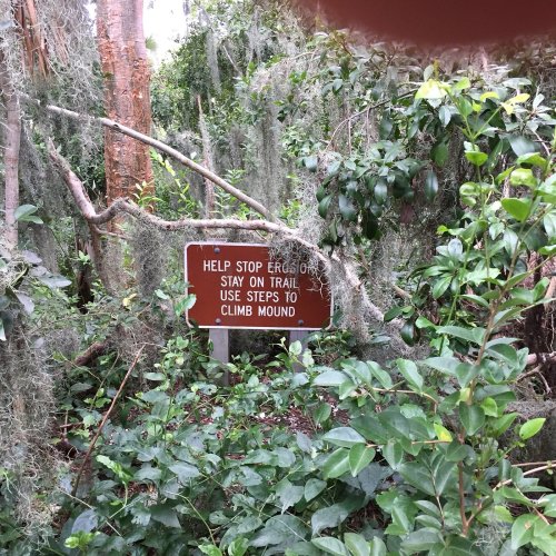A sign in a wooded area reads: "Help stop erosion. Stay on trail. Use steps to climb mound." Surrounded by greenery.