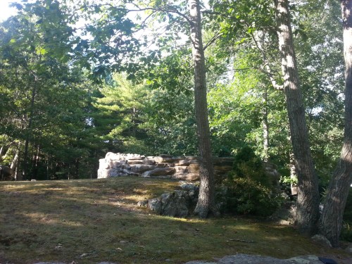 A stone structure surrounded by trees in a lush, green forest setting. Sunlight filters through the leaves.