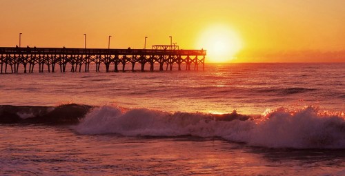 A vibrant sunset over the ocean, with waves crashing and a pier silhouetted against the colorful sky.