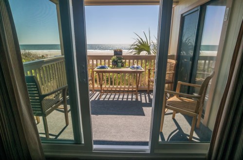 A view from inside a room looking out to a sunny balcony with a table and chairs overlooking the ocean.