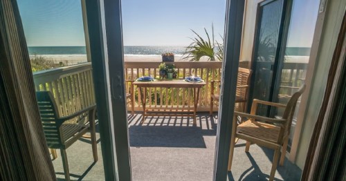 A sunny balcony with a table and chairs overlooking a beach and ocean view.