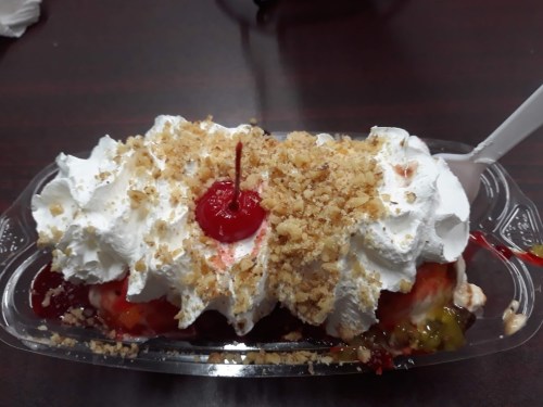 A colorful dessert topped with whipped cream, a cherry, and crushed nuts in a clear plastic dish.