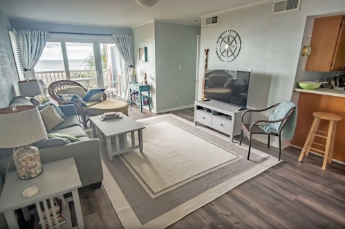 Cozy living room with a view, featuring a sofa, TV, and light decor, leading to a balcony with ocean scenery.