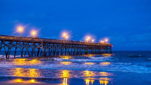A wooden pier illuminated by lights at dusk, reflecting on calm ocean waves under a blue sky.