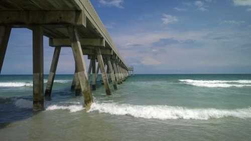 A wooden pier extends over calm waves, with a clear blue sky and gentle clouds in the background.