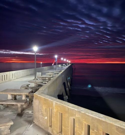 A pier extends into the ocean at sunset, with glowing lamps and dramatic clouds in the sky.