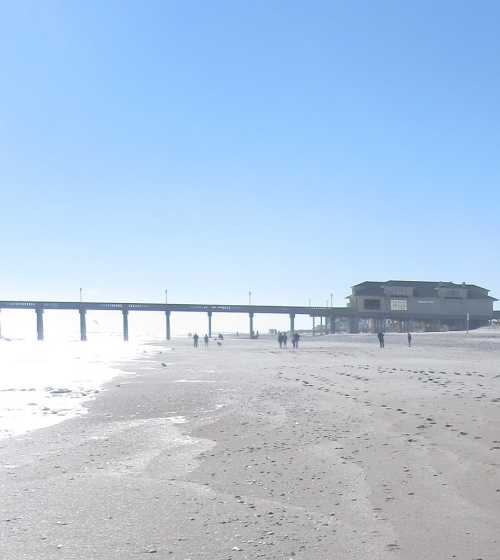 A sunny beach scene with a pier in the distance and people walking along the shore. Waves gently lap at the sand.