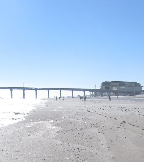 A sunny beach scene with a pier in the distance and people walking along the shore. Waves gently lap at the sand.