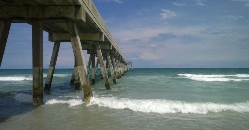 A wooden pier extends over calm blue waters, with gentle waves lapping at its supports under a cloudy sky.