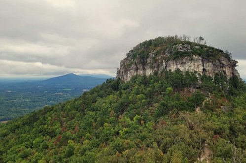A rocky mountain peak surrounded by lush greenery under a cloudy sky, with distant hills in the background.