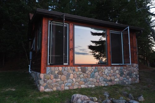 A small cabin with large windows reflecting a sunset, surrounded by trees and a stone foundation.