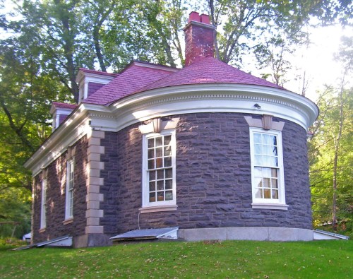 A round stone house with a red roof, surrounded by trees and grass, featuring large windows and a chimney.