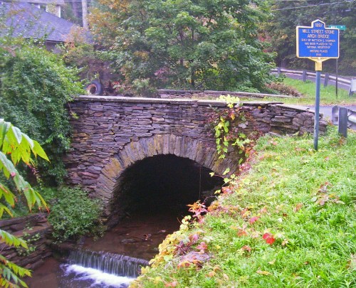 Stone arch bridge over a small stream, surrounded by greenery and a historical marker nearby.