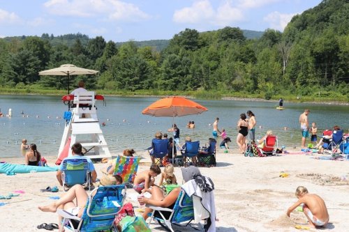 A busy beach scene with people swimming, lounging on chairs, and a lifeguard watching over a lake under a sunny sky.