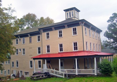 Historic three-story building with a red roof, white porch, and a cupola, surrounded by trees and grassy area.