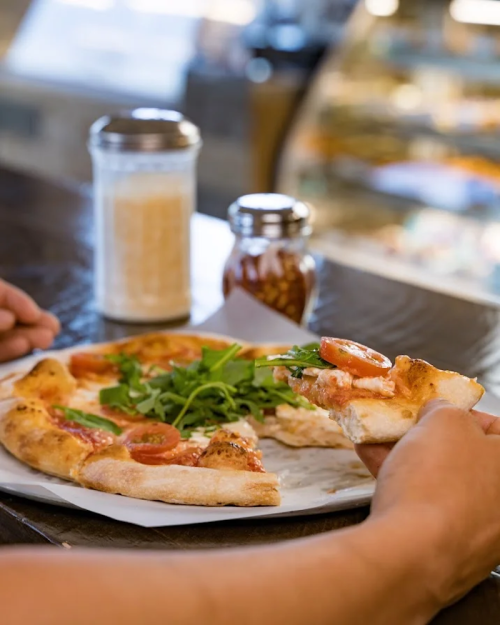 A person holds a slice of pizza topped with tomatoes and arugula, with a drink and condiments in the background.