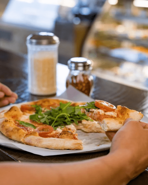 A person holds a slice of pizza topped with tomatoes and arugula, with a drink and condiments in the background.