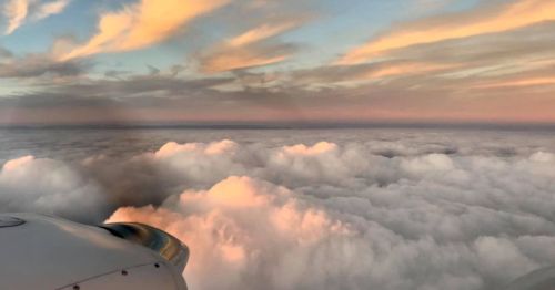 Aerial view of fluffy clouds at sunset, with soft pastel colors in the sky and a glimpse of an airplane wing.