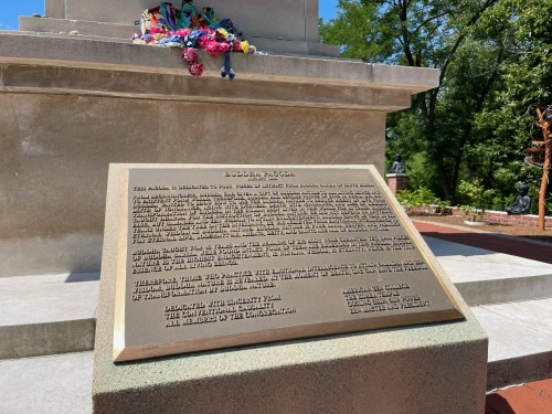 A bronze plaque on a monument, surrounded by flowers, detailing historical information and dedication.