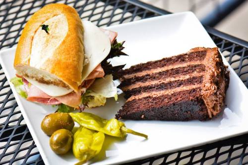 A plate with a sandwich stacked with meats and cheese, a slice of chocolate cake, and pickles on the side.