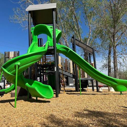 A vibrant green playground slide set against a clear blue sky, surrounded by trees and wood chips.