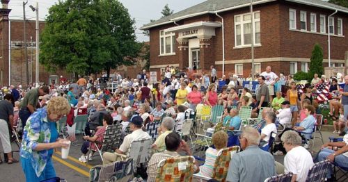 A large crowd gathers outside a library, seated in folding chairs, enjoying a community event on a sunny day.