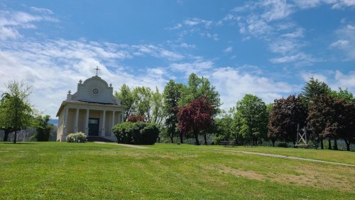 A small chapel surrounded by lush greenery and trees under a bright blue sky with scattered clouds.
