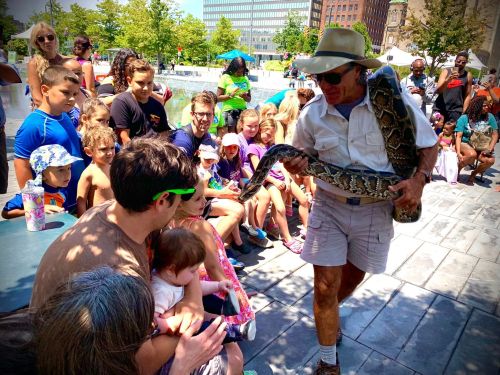 A man in a hat holds a snake while engaging with a crowd of children and adults at an outdoor event.