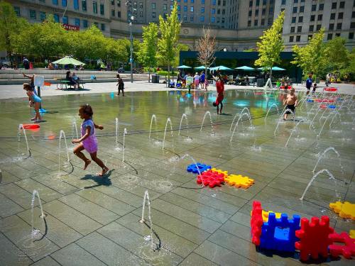 Children play in a splash pad, running through water jets and colorful foam blocks on a sunny day.