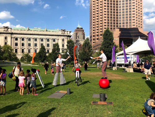 Two performers in striped outfits juggle and balance on a ball, while a crowd watches in a park with city buildings in the background.