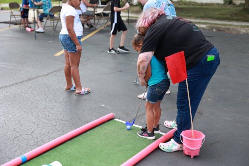 A child is guided by an adult while putting a golf ball on a mini-golf course, with others playing in the background.
