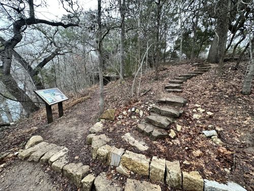 A winding stone path leads through a wooded area, with a signpost nearby and fallen leaves covering the ground.