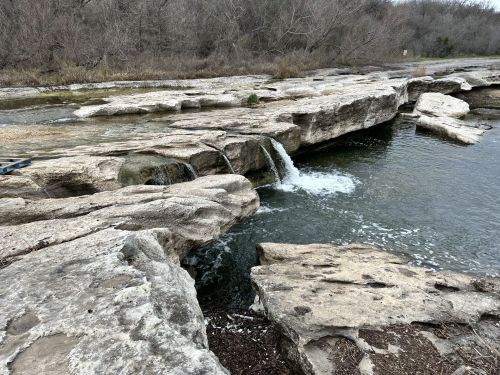 A rocky riverbank with a small waterfall flowing into a calm pool, surrounded by bare trees and a serene landscape.