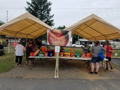 A busy outdoor raffle booth at a Peach Festival, with colorful containers and people gathered under tents.