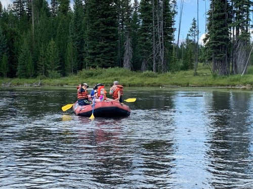 A group of people in a red raft paddling on a calm river surrounded by lush green trees and a clear blue sky.