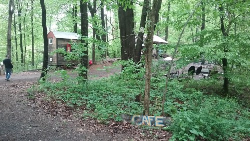 A wooded area with a small café sign, surrounded by greenery and rustic buildings in the background.