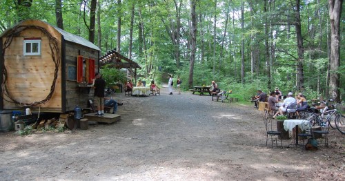 A rustic outdoor café in a wooded area, with people dining, biking, and enjoying nature.