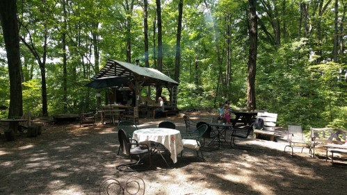 A rustic outdoor gathering area with tables and chairs surrounded by trees, featuring a small shelter and smoke rising.