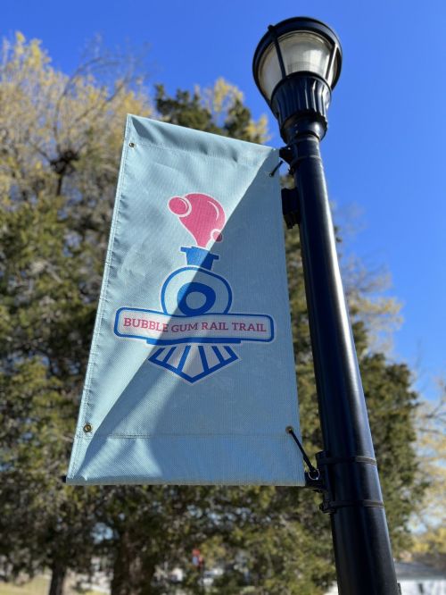 A banner for the Bubble Gum Rail Trail featuring a train graphic, hanging from a lamp post against a clear blue sky.