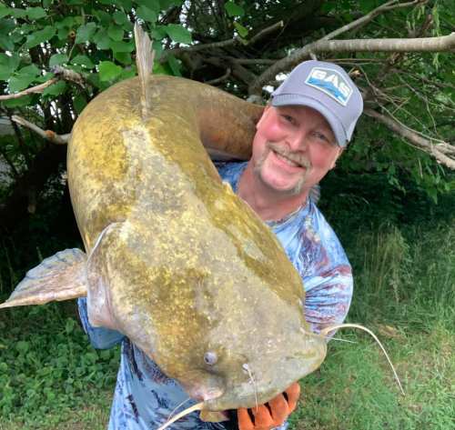 A smiling person holds a large catfish in a natural outdoor setting, surrounded by trees and greenery.