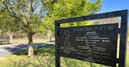 Sign for the Bubble Gum Rail Trail, listing advocates and sponsors, with trees and a path in the background.