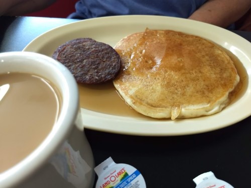 A plate with a large pancake and a sausage patty, accompanied by a cup of coffee and creamers.