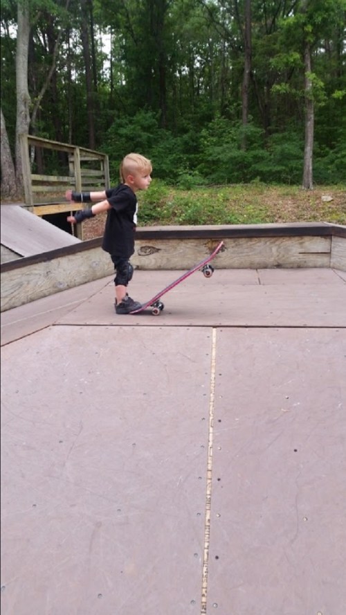 A young child skateboards on a ramp in a park, wearing gloves and a black shirt, surrounded by trees.