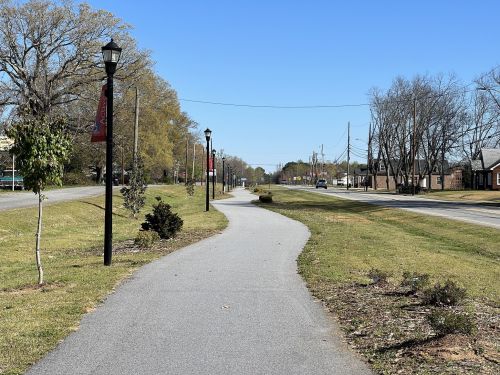 A paved pathway lined with lampposts, leading through grassy areas alongside a road under a clear blue sky.