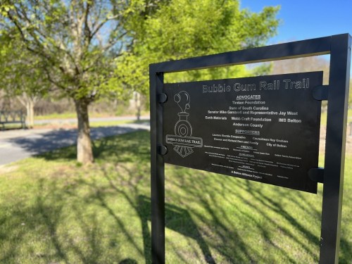 Sign for the Bubble Gum Rail Trail, featuring names of advocates and sponsors, set against a green landscape.