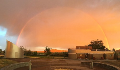 A vibrant rainbow arcs over a building at sunset, with orange and pink hues in the sky and greenery in the background.