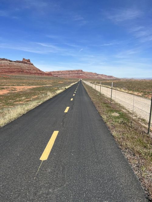 A long, empty road stretches into the distance, flanked by red rock formations and a clear blue sky.