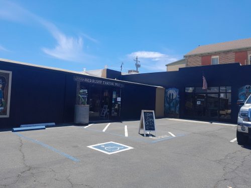 A storefront with a black exterior, featuring signs for "Red Bluff Trading Post" and an open sign, parked cars in front.